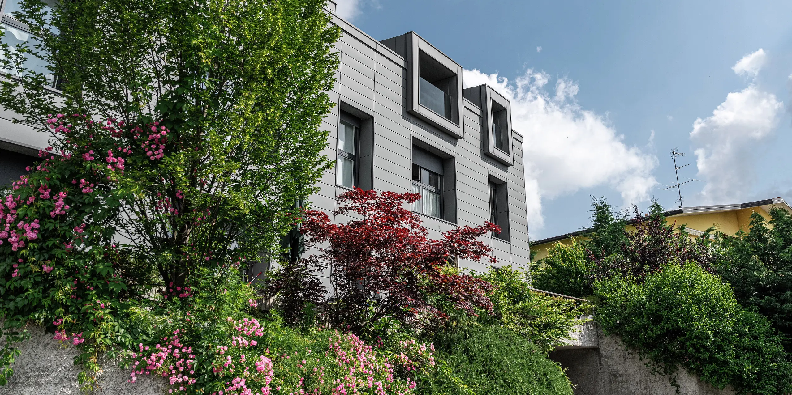 Moderne boligkompleks i Faloppio, Italia, med stilfull fasade av PREFA Sidings. De utkragende vinduene er et unikt arkitektonisk blikkfang, mens plantene og blomstene rundt understreker bygningens naturlige skjønnhet. Den holdbare aluminiumsbekledningen fra PREFA er en estetisk og funksjonell løsning i all slags vær.