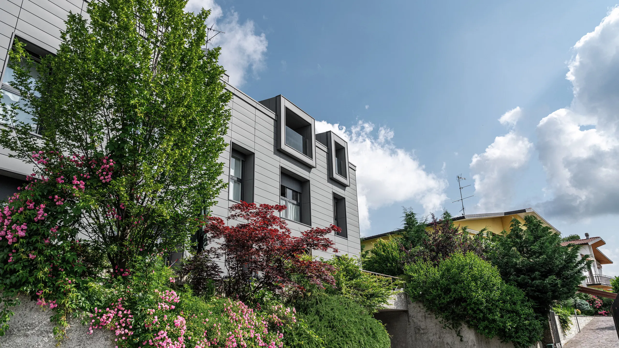 Moderne boligkompleks i Faloppio, Italia, med stilfull fasade av PREFA Sidings. De utkragende vinduene er et unikt arkitektonisk blikkfang, mens plantene og blomstene rundt understreker bygningens naturlige skjønnhet. Den holdbare aluminiumsbekledningen fra PREFA er en estetisk og funksjonell løsning i all slags vær.