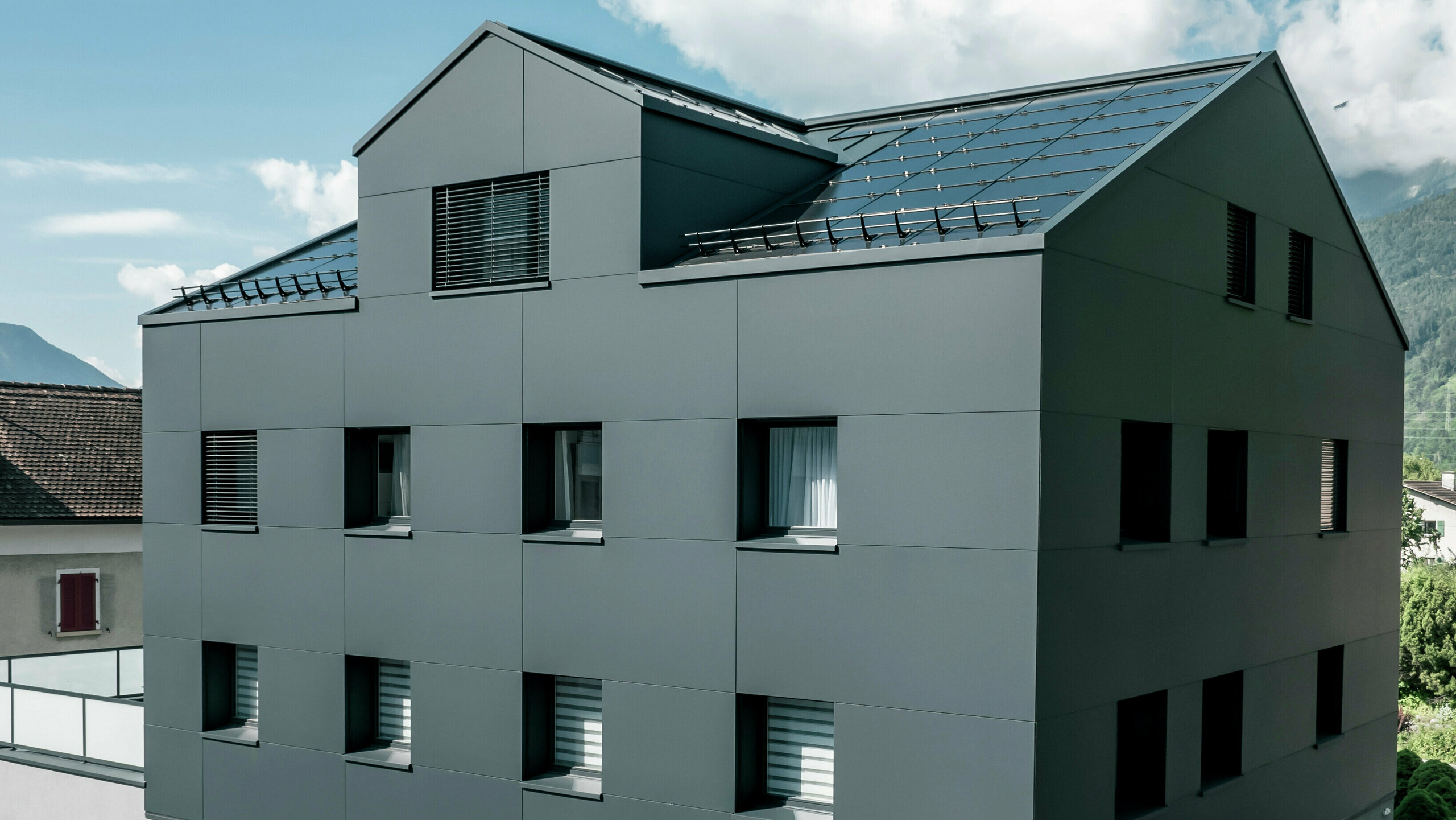 Modernisiertes Wohn- und Geschäftsgebäude nach der Fassadensanierung mit PREFABOND Aluminium Verbundplatten in Anthrazit. Die klare, elegante Linienführung der neuen Fassade verleiht dem Gebäude ein modernes Erscheinungsbild. Im Erdgeschoss befindet sich das Architekturbüro Voser. Die Umgebung mit Bäumen und Bergen rundet die harmonische Optik ab.