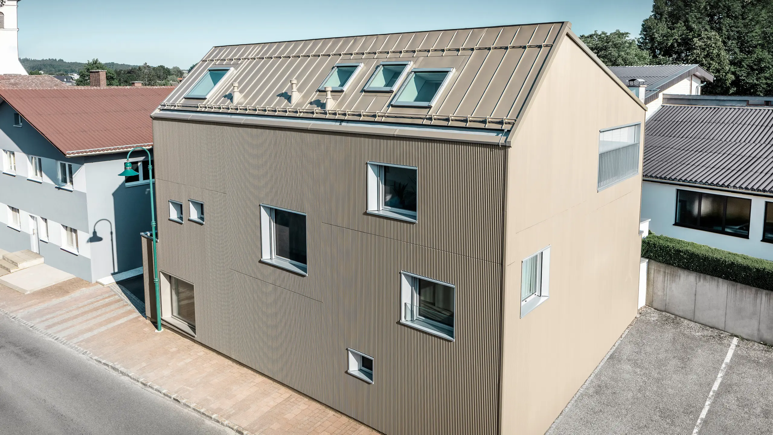Fugleperspektiv på en enebolig i Frankenburg, Østerrike, som skiller seg ut med sin PREFA-fasade i form av et sikksakkprofil og et PREFALZ-tak, begge i en matchende bronsefarge. Fasaden har en markant tekstur som fanger opp det naturlige lyset og skaper et levende skygge-spill. Flere takvinduer slipper naturlig lys innvendig og understreker den funksjonelle og estetiske integreringen av moderne arkitektoniske elementer. Huset passer harmonisk inn i det typiske østerrikske landsbymiljøet med sitt moderne design, samtidig som det skaper en kontrast til de omkringliggende tradisjonelle bygningene.