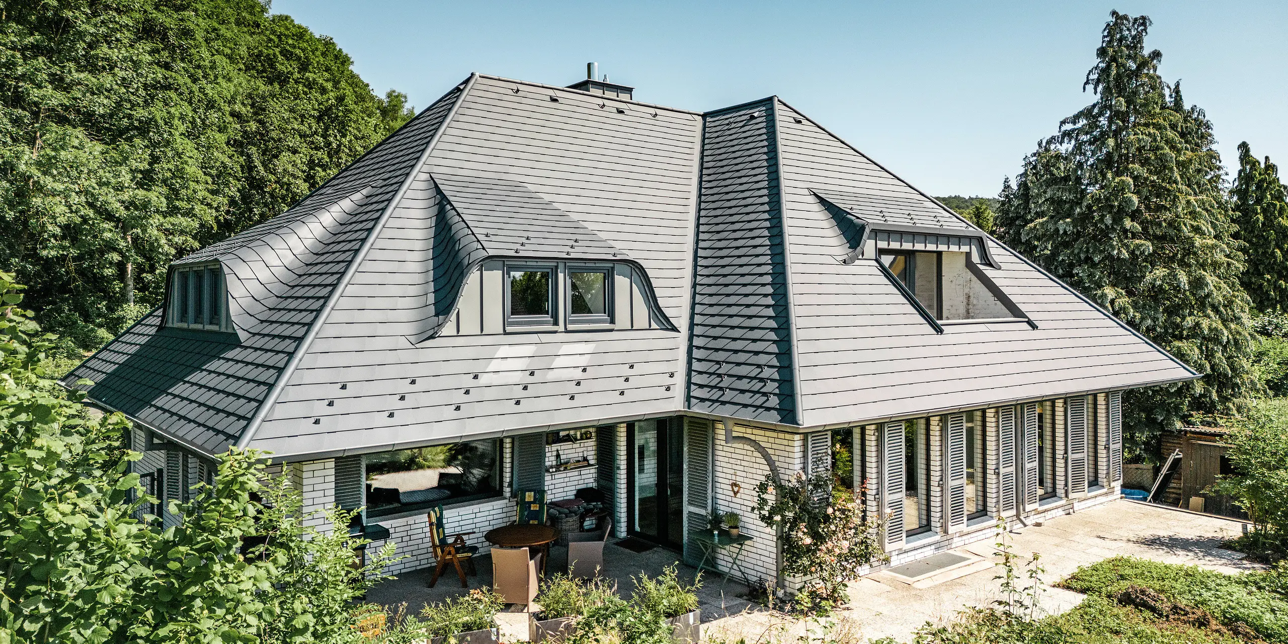 Moderne enebolig i Einbeck, Tyskland, med PREFA takspon i P.10 antrasitt. De holdbare PREFA taksponene i aluminium er kjent for sin kvalitet og holdbarhet. Takkonstruksjonen på bygningen understreker den individuelle arkitekturen med takvinduer, som fremheves av fleksibiliteten til PREFA taksponene. Takavløpet i form av PREFA avløpsrør og takrenne er også i antrasitt. Rundt eneboligen er det en stor og velstelt hage og en terrasse. Bak huset ligger en skog som sammen med den blå himmelen skaper en naturlig atmosfære.