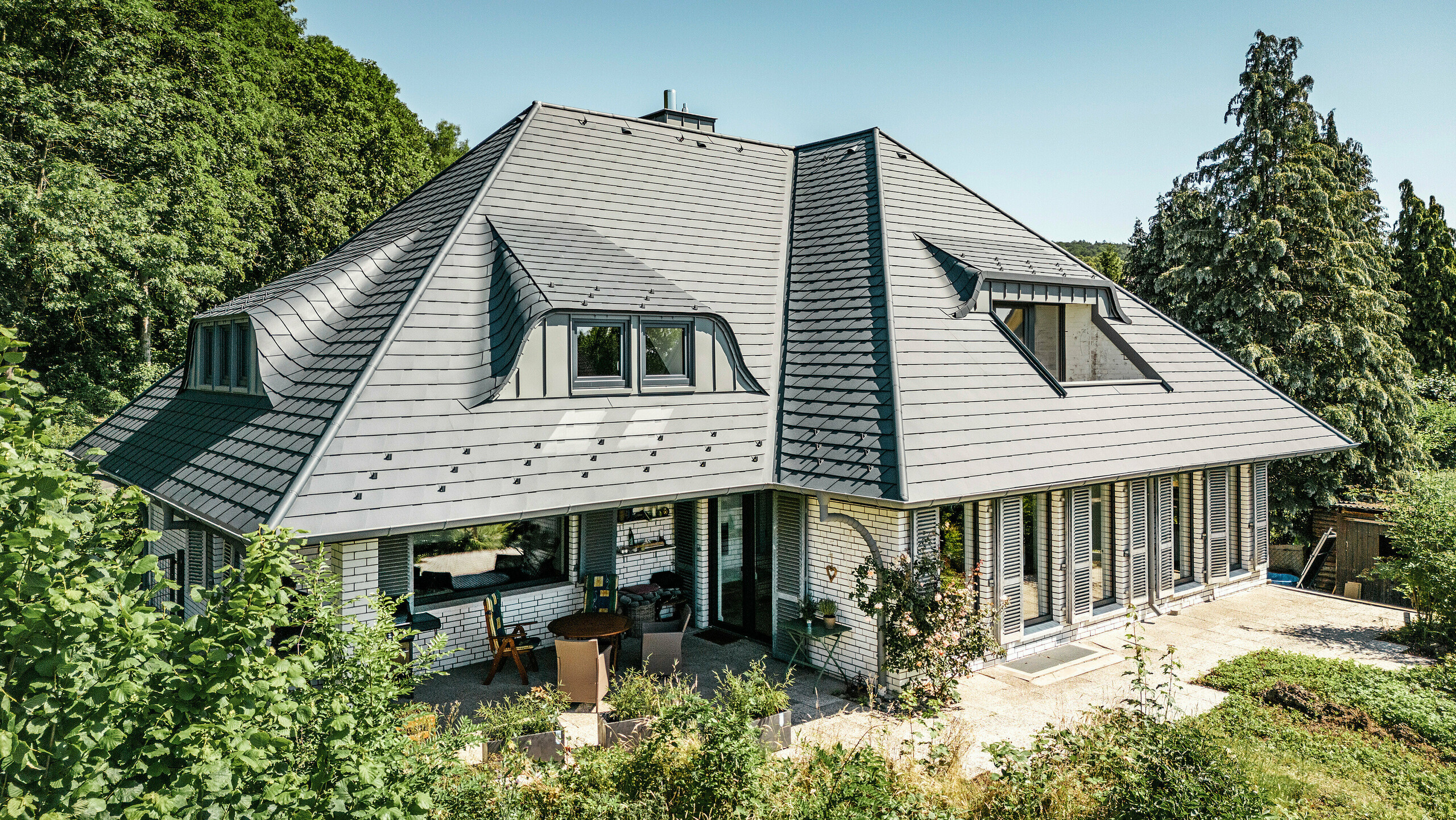 Moderne enebolig i Einbeck, Tyskland, med PREFA takspon i P.10 antrasitt. De holdbare PREFA taksponene i aluminium er kjent for sin kvalitet og holdbarhet. Takkonstruksjonen på bygningen understreker den individuelle arkitekturen med takvinduer, som fremheves av fleksibiliteten til PREFA taksponene. Takavløpet i form av PREFA avløpsrør og takrenne er også i antrasitt. Rundt eneboligen er det en stor og velstelt hage og en terrasse. Bak huset ligger en skog som sammen med den blå himmelen skaper en naturlig atmosfære.