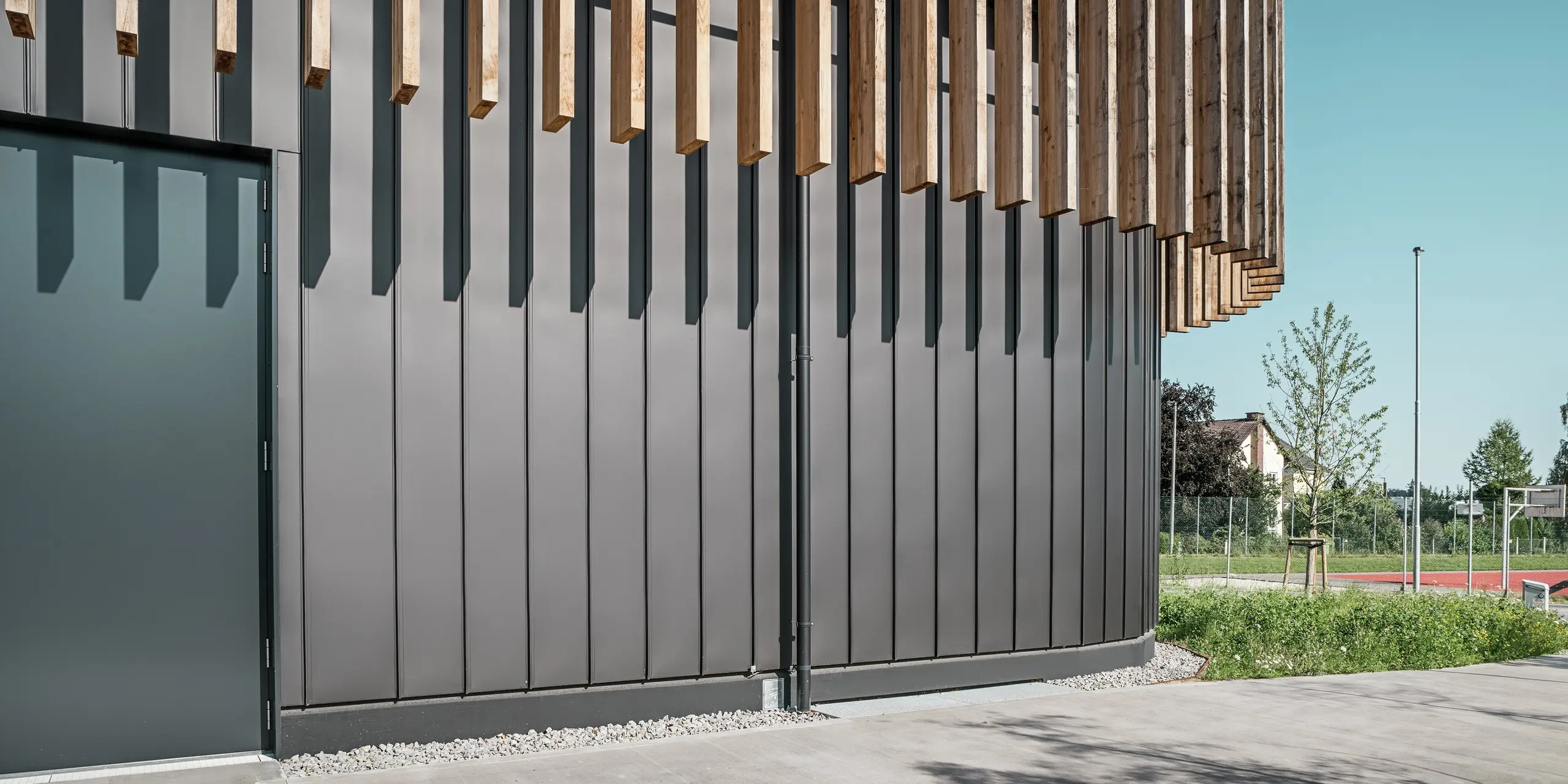 Nahaufnahme des Erdgeschosses der Team 7 Welt in Ried im Innkreis: Die schwarzbraune PREFALZ Fassade bildet mit den vertikalen Holzelementen ein harmonisches Gesamtbild. Die robuste Aluminiumverkleidung schützt vor Witterungseinflüssen und überzeugt durch ihre langlebige, wartungsfreie Qualität. Das Spiel von Licht und Schatten unterstreicht die klaren Linien und die moderne Architektur des Gebäudekomplexes.