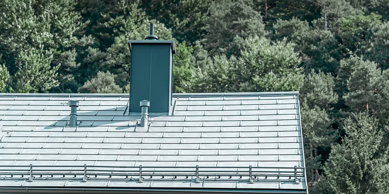 Detaljvisning av et saltak, kledd med PREFA takplate R.16 i steingrå med snøfangersystem, pipebeslag, takrenne og ventilasjonsrør, også fra PREFA