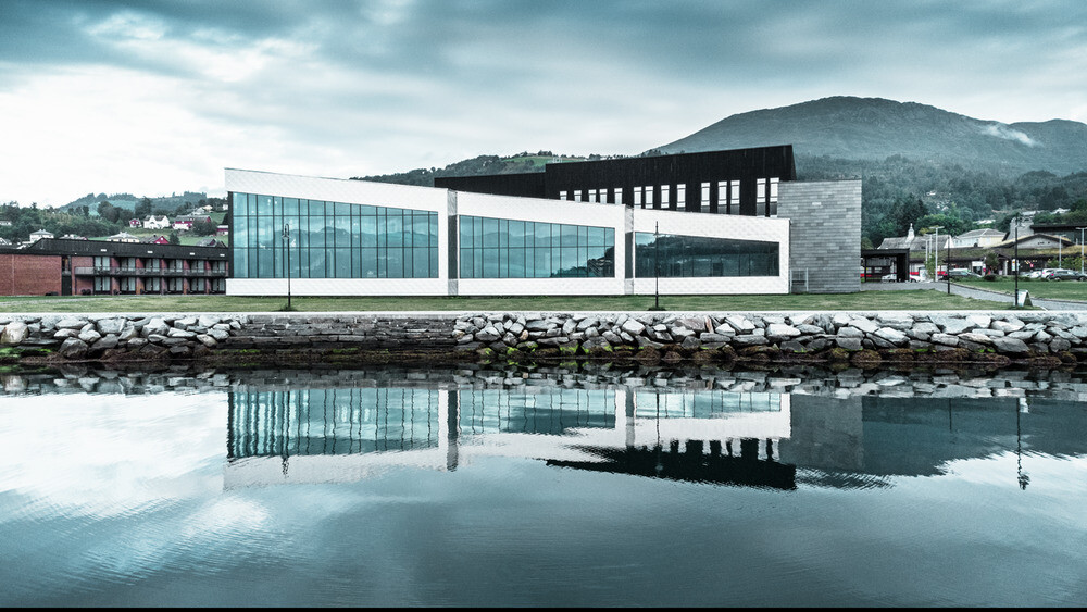 In der Abbildung ist das Healthcare Center in Oystese vom Wasser aus zu sehen. Diese spiegelt sich im Wasser des Hardangerfjord und wurde mit der Wandraute 44x44 in Naturblank eingedeckt. Der Himmel dahinter ist Wolkenbedeckt.