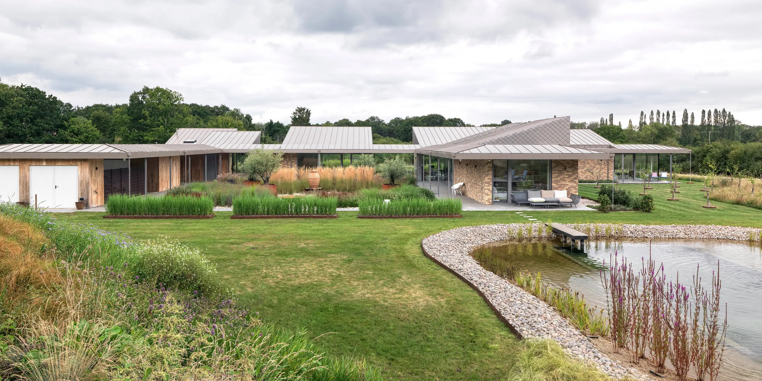 Weitwinkelansicht des architektonisch anspruchsvollen Wohnhauses in West Sussex mit großflächigem PREFALZ Aluminiumdach in P.10 Bronze und harmonisch integrierten PREFA Wandrauten in ausgewählten Fassadenbereichen. Das moderne Gebäude erstreckt sich flach und weitläufig über das Grundstück, eingebettet in eine naturnahe Bepflanzung und mit einem gestalteten Naturpool im Vordergrund. Die robusten PREFA Produkte unterstützen das nachhaltige Baukonzept und sorgen für ein einheitliches, langlebiges Erscheinungsbild.