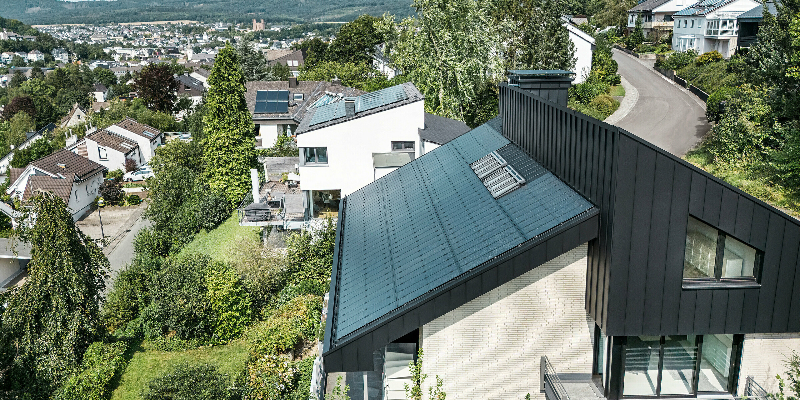 Panoramaansicht eines Einfamilienhauses in Meschede, Deutschland, mit einem versetzten Pultdach aus PREFALZ in P.10 Schwarz. Das Dach ist mit rahmenlosen Glas/Glas-PV-Modulen von PREFA ausgestattet, die Nachhaltigkeit und Design vereinen. Die Aluminiumverkleidung der Fassade am Giebel ergänzt die klaren Linien und die moderne Architektur, eingebettet in eine grüne Wohnsiedlung mit Blick auf die umliegende Landschaft und Stadt.