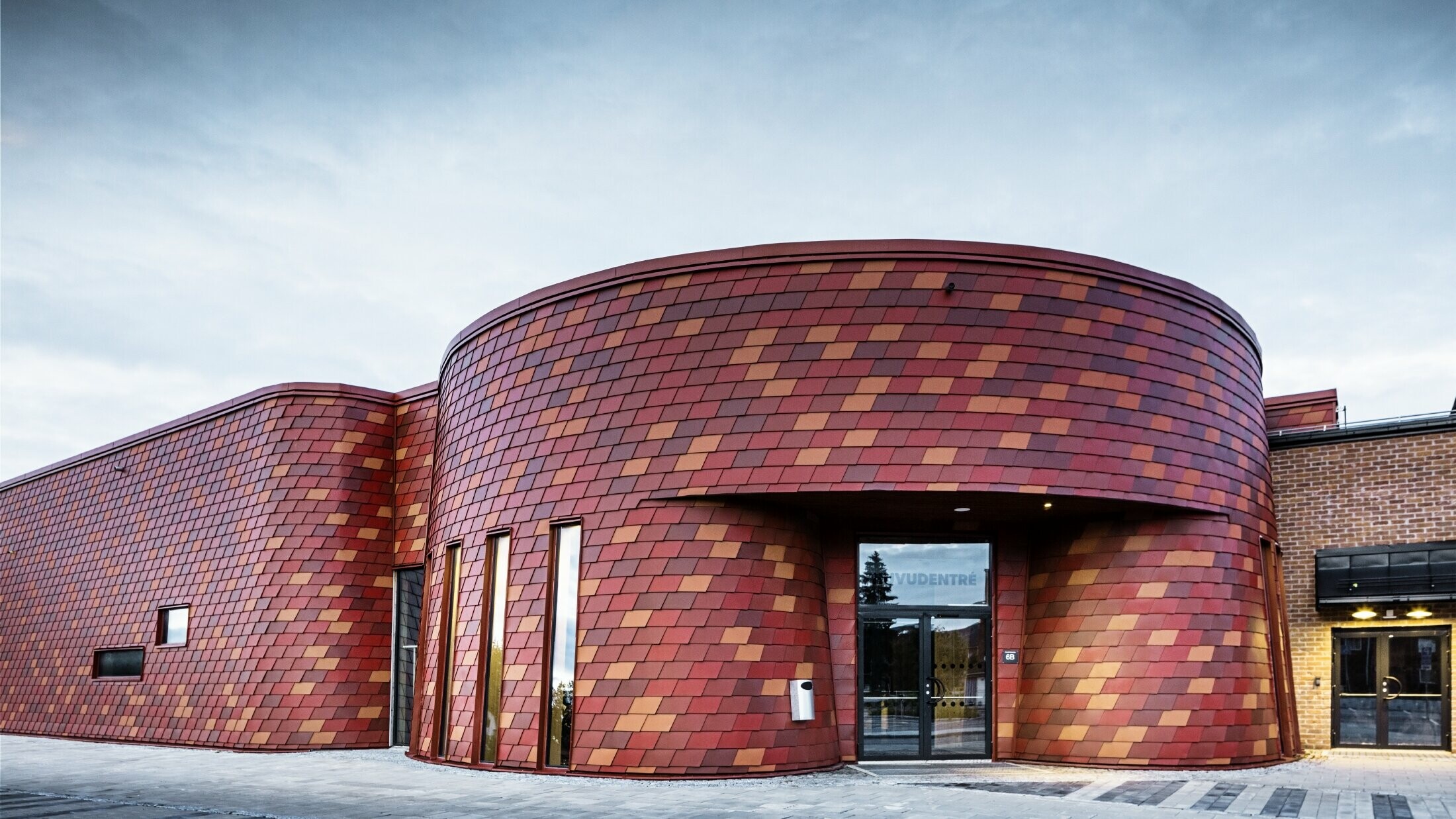 Ishall med flatt tak og avrundet form, fasaden er kledd med veggshingel i aluminium i forskjellige rødtoner fra PREFA - oksidrød, mursteinsrød, rødbrun, brunrød
