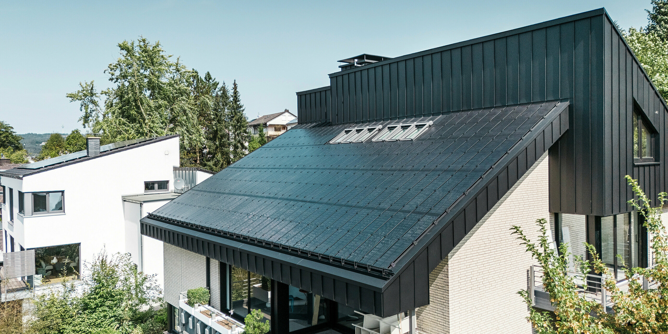 Moderne Architektur eines Einfamilienhauses in Meschede, Deutschland, mit einem PREFALZ Aluminiumdach in P.10 Schwarz und integrierten Glas/Glas PV-Modulen. Das versetzte Pultdach verbindet zeitloses Design mit nachhaltiger Photovoltaik-Technologie. Die innovativen PREFALZ Solarmodule liefern eine Leistung von 18 kWp. Die rahmenlosen Module und die elegante Aluminiumverkleidung von Fassade und Dach bilden eine ästhetische und funktionale Einheit, die sich perfekt in das grüne Wohnumfeld einfügt.