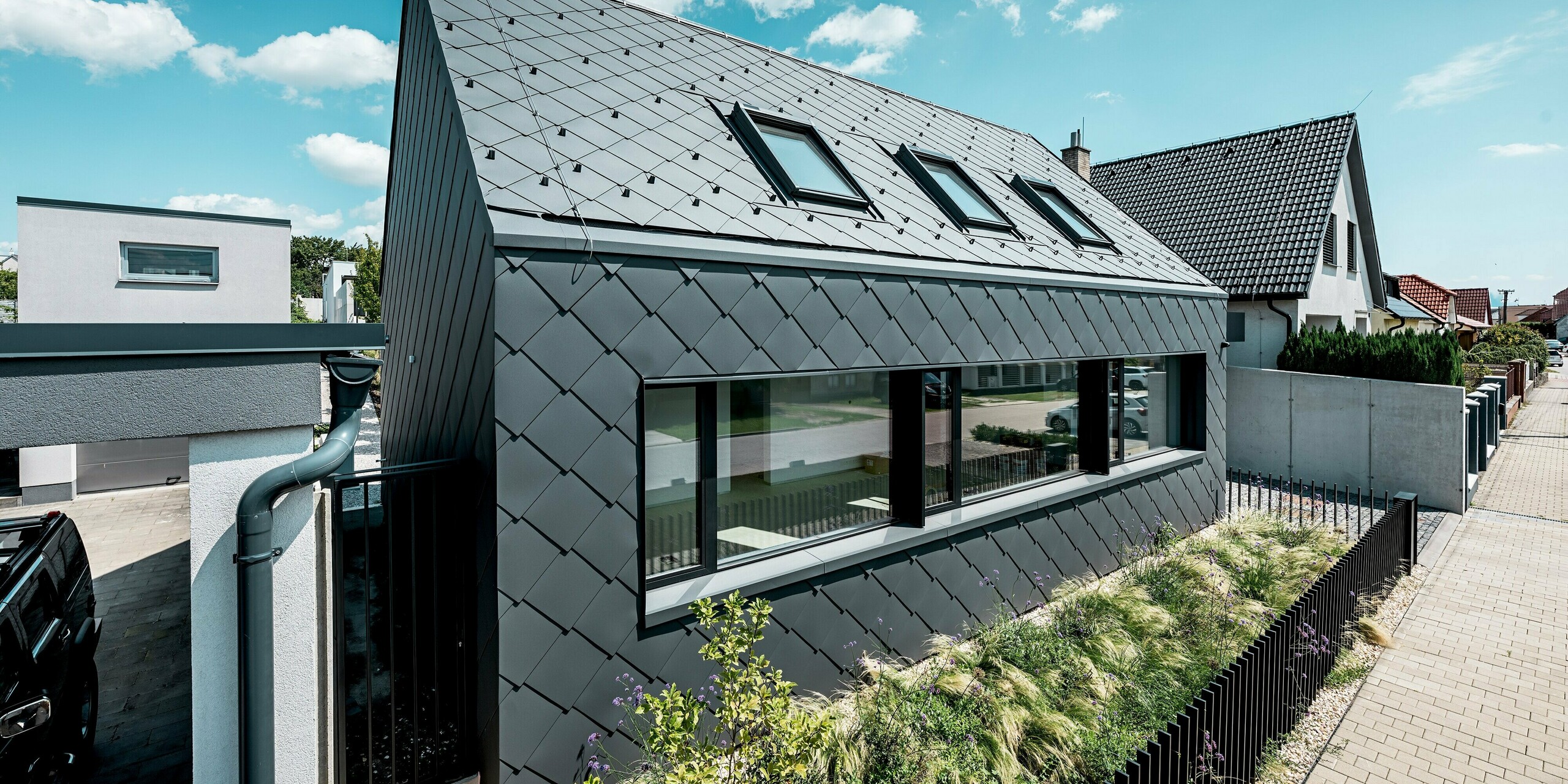 Das Bild zeigt die moderne Vorderseite des Einfamilienhauses in Trnava-Modranka aus schräger Perspektive. Das Haus wurde mit PREFA Dach- und Wandrauten 44 × 44 in der Farbe P.10 Dunkelgrau vollständig verkleidet. Die durchgehende Rautenstruktur erzeugt eine harmonische, hochwertige Gebäudehülle, während die großen Fensterflächen für eine klare, zeitgemäße Optik sorgen. Die robusten PREFA-Aluminiumprodukte bieten dauerhaften Schutz vor Witterungseinflüssen, sind formstabil und haben farbbeständige Oberflächen. Der blaue Himmel und die gepflegte Begrünung unterstreichen den prägnanten Charakter des modernen Baukörpers.